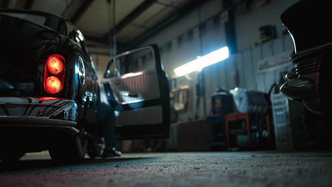 Vintage black car with its driver's door open inside a dimly lit auto repair garage under a single overhead shop light