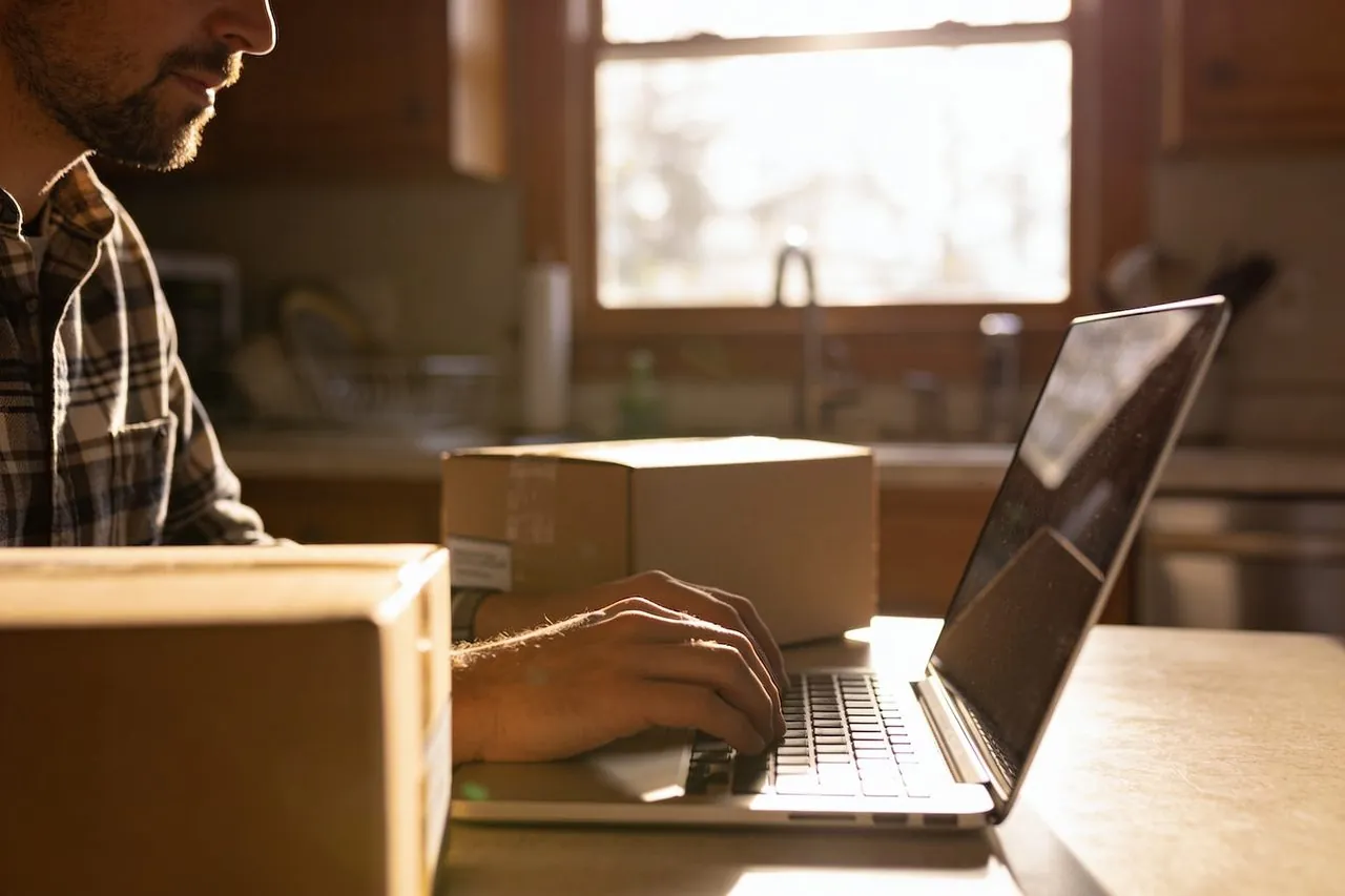 Home-based business owner typing on a laptop beside shipping boxes at a kitchen table