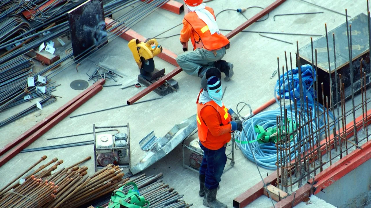 Two construction workers in orange safety vests on a concrete deck surrounded by rebar bundles, a cut-off saw, portable generators and coiled hoses