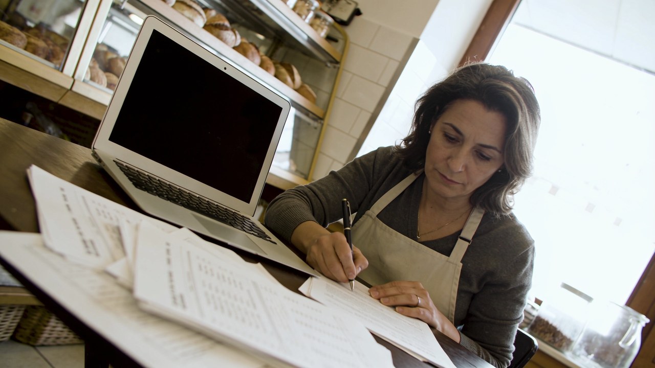 Bakery owner in an apron sitting at a back-office table writing on financing paperwork spread beside an open laptop in morning light