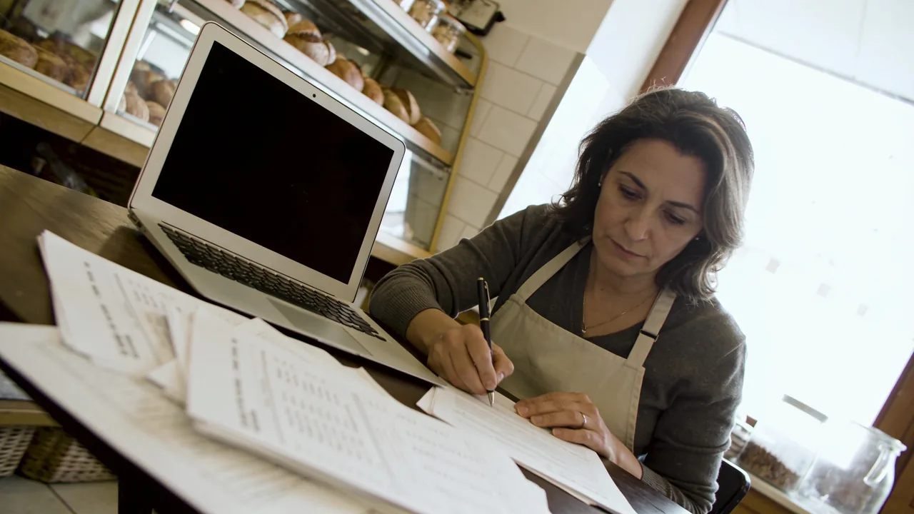 Bakery owner in an apron sitting at a back-office table writing on financing paperwork spread beside an open laptop in morning light