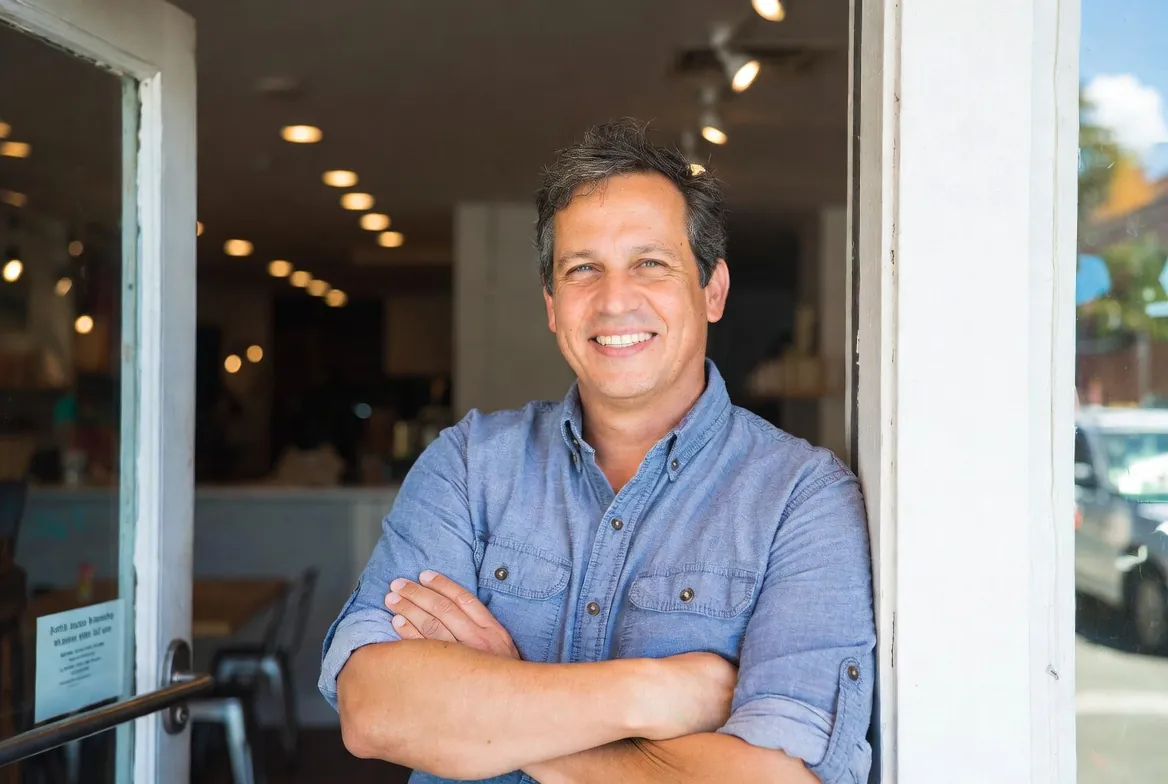 Small business owner standing in the doorway of his shop, arms crossed and smiling — the kind of customer The Broker Shop funds every day